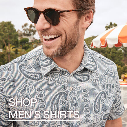 A close up image of a man wearing a light blue polo with a paisley print in dark blue. He is wearing sunglasses and the words Shop Men's Shirts read across the bottom left of the image. 